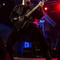 A musician with a beard, wearing a black outfit, passionately plays an electric guitar on a dimly lit stage with vibrant red and purple lights. The stage has metal structures and amplifiers in the background. The musician stands in a wide stance, fully immersed in the performance at Kilkim Žaibu 2015.