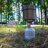 A portable camping stove with a small fuel canister heats a pot with a ladle inside. The stove is set on a grassy and wooded area with several colorful tents visible in the background, reminiscent of a lively 2015 Kilkim Žaibu festival. The scene is bright and sunny, with tall trees providing a green canopy.