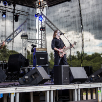 At Kilkim Žaibu festival in 2015, four musicians dressed in black, with painted faces, perform on an outdoor stage. Two play electric guitars, one plays bass, and the other plays drums. The stage features a metal frame, lighting equipment, and speakers. Trees are visible in the background. The sky is overcast.