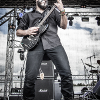 A guitarist with face paint performs passionately on stage at the 2015 Kilkim Žaibu festival. He wears black clothing and combat boots, playing an electric guitar. The background features large Marshall amplifiers and stage lighting equipment against a partially cloudy sky.