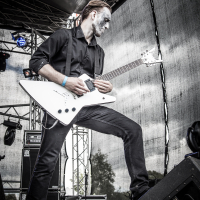 A musician with white face paint is playing a white electric guitar on an outdoor stage at the 2015 Kilkim Žaibu festival. He's dressed in black, including a black shirt and pants, and stands in a powerful stance with one foot on an amplifier. The background includes stage equipment and a mesh backdrop filtering daylight.