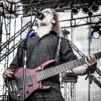 At the 2015 Kilkim Žaibu festival, a musician with long hair passionately sings into a microphone while playing a six-string bass guitar on an outdoor stage. He is wearing a black shirt and has several wristbands on his right wrist, framed by the stage's scaffolds and lighting equipment in the background.