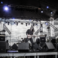A rock band performs energetically on an outdoor stage at the 2015 Kilkim Žaibu, surrounded by musical equipment and illuminated by blue and white stage lights. Four musicians play electric guitars and bass, with a drum set visible in the background. They are under a large, metal-framed canopy.