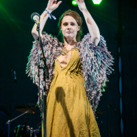 A woman stands on stage illuminated by green stage lights at the 2015 Kilkim Žaibu festival. She wears a flowing, mustard yellow dress and a shawl with feathery textures. Her arms are raised, and she stands in front of a microphone. The background shows part of a drum set, and the atmosphere suggests a live music performance.