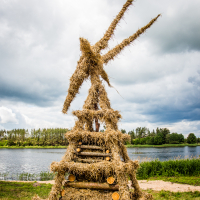 A large, artistic sculpture made of hay and logs stands on grassy ground near a lake. The structure is pyramid-shaped with triangular extensions at the top, resembling a figure holding spears. Created in 2015 for the Kilkim Žaibu festival, it sits under an overcast sky with trees lining the opposite bank of the lake.