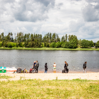 A group of people stands on a sandy beach beside a calm lake, with lush green trees lining the opposite shore. Some individuals are near the water while others are on the sand. Several paddle boats are on the shore, and the sky above is partly cloudy with patches of blue, reminiscent of Kilkim Žaibu 2015.