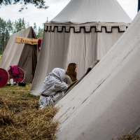 People in medieval-style clothing sit and interact near large, beige canvas tents at an outdoor camp setup during Kilkim Žaibu 2015. Two red wooden shields lean against one tent in the background, and a wooden sign with illegible text hangs nearby. The area is surrounded by trees and covered with straw.