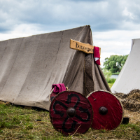 A medieval-themed camp scene from Kilkim Žaibu 2015 features a canvas tent with a wooden sign above its entrance. Two painted wooden shields, one red with a black pattern, lean against the tent. Another similar tent and a pile of hay are in the background, with a river and trees visible under a cloudy sky.