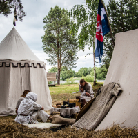 A scene at the 2015 Kilkim Žaibu outdoor historical reenactment camp shows two canvas tents set up among trees. Two people in period clothing sit near a table with wooden items and food. A river, flags, and more trees are in the background. The atmosphere is rustic and tranquil.
