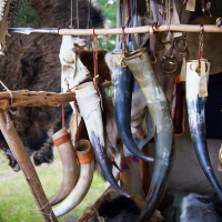 A display of assorted animal horns hangs from a wooden structure at an outdoor market, reminiscent of Kilkim Žaibu 2015. The horns vary in size and color, adorned with leather straps and metal rings. In the background, fur pelts and other traditional items create a rustic, historical ambiance.
