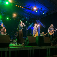 A band performs on an illuminated stage at Kilkim Žaibu 2015, with green, blue, and yellow lights. Six musicians—two vocalists, a guitarist, a bassist, a drummer, and a keyboardist—shine against the metal trusses and dark backdrop at this vibrant music festival.