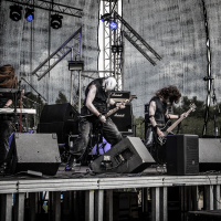 At the 2015 Kilkim Žaibu festival, a heavy metal band performs energetically onstage. Members include a keyboardist, two guitarists, and a drummer in the background. Amplifiers and speakers surround the stage. Intense lighting and a dark backdrop add to the moody, powerful atmosphere of the outdoor concert.