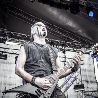 A musician passionately performs on stage at the 2015 Kilkim Žaibu event, playing an electric guitar. He wears black clothing and face paint that gives him a dramatic look. The background displays stage lighting and equipment, enhancing the energetic atmosphere of the live concert.