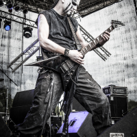 A musician with face paint, wearing dark, rugged attire, plays an electric guitar passionately on an outdoor stage at the 2015 Kilkim Žaibu festival. Bright stage lights illuminate him from above, and various stage equipment is visible. The background consists of metal scaffolding and a draped, semi-transparent tarp.