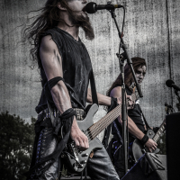 Two musicians perform energetically on stage at Kilkim Žaibu 2015, adorned in black metal attire with painted white and black faces. One sings into a microphone while playing a bass guitar, while the other strums an electric guitar. The backdrop is a gray, textured wall and trees are visible in the distance.