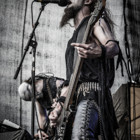 At the 2015 Kilkim Žaibu festival, a guitarist passionately performs on stage wearing black leather pants, a sleeveless shirt, wristbands, and face paint. The musician, with long hair and a beard, sings into a microphone while another band member plays guitar in the background. The setting is a live concert with a gritty, dark aesthetic.