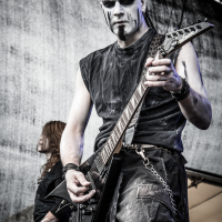 A guitarist with black-and-white face paint and black sleeveless attire performs on stage at the 2015 Kilkim Žaibu festival, gripping a black electric guitar. Background: a partially visible band member, drums, and stage equipment. The scene is set outdoors with a gray backdrop. A website watermark, "xlitephoto.net," is at the bottom right.