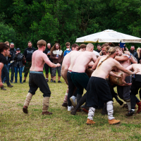 A group of shirtless people are participating in a wrestling match on grassy ground at the Kilkim Žaibu festival in 2015. Spectators, some dressed in casual attire while others wear themed costumes, stand around watching. A canopy umbrella is visible in the background amidst a green wooded area.