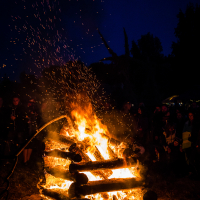 At the Kilkim Žaibu festival in 2015, a large bonfire burns brightly at night, with flames and sparks rising upwards. People are gathered around the fire, silhouetted against its light. The sky is dark blue, and a bright moon is visible above. Sparse trees and additional people can be seen in the background.
