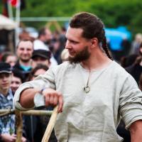 A man dressed in medieval attire stands outdoors at the 2015 Kilkim Žaibu festival, holding a wooden staff. He has long hair braided on one side, a beard, and is wearing a beige tunic with a decorative brooch and a woven belt. In the background, a crowd of people are watching, and there are trees and banners visible.