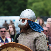 A person dressed in medieval armor stands in front of a blurred outdoor crowd at the 2015 Kilkim Žaibu festival. They wear a metal helmet with a protective face covering and a quilted garment with blue fabric lining at the neck, holding a large round wooden shield. Forested greenery is visible in the background.