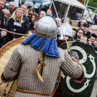 Two people in medieval armor, one with a long blonde braid, metal helmet, padded jacket, and shield, face each other in combat reenactment. A crowd at the 2015 Kilkim Žaibu festival watches intently from behind a rope barrier at the outdoor event. Tents and more spectators are visible in the background.