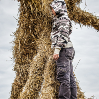 A person wearing a camouflage hoodie and dark cargo pants stands on a large, straw-covered structure at the 2015 Kilkim Žaibu festival. Their head is turned away, facing the textured straw surface, as an overcast sky looms in the background.