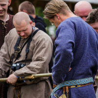 A group of men in medieval attire participates in an outdoor historical reenactment at the 2015 Kilkim Žaibu festival. The man in the foreground wears a blue tunic with a decorative belt, while the man to his left, in a brown tunic, holds a sword and has a camera around his neck. Other participants are partly visible in the background.