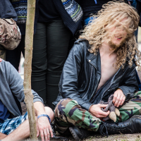 At the 2015 Kilkim Žaibu festival, a crowd is gathered outdoors. In the foreground, a person with long, curly blonde hair and a black leather jacket sits on the ground with legs crossed, wearing camouflage pants and black boots. To the left, another person wearing a gray hoodie and plaid shorts is sitting and smiling at them.