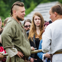 At the Kilkim Žaibu festival in 2015, a man with a short blonde mohawk, beard, and traditional green attire stands in a crowd, facing another man wearing a white robe and holding a microphone. Ornamental items hang from their belts. Onlookers, including a woman with long red hair, observe the scene against a blurry green background.