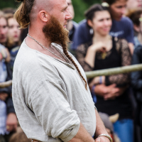 A man with a long beard and a partially shaved head, the remaining hair tied in a top knot, stands outdoors at the 2015 Kilkim Žaibu festival. He wears a loose, light-colored shirt and a woven belt with pouches. A crowd of people watches intently in the background.