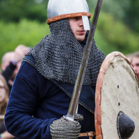 A person dressed in medieval armor holds a sword and a round shield. They wear a chainmail coif with a steel helmet and full medieval attire. The background shows an outdoor setting with blurred greenery and several people watching, reminiscent of the 2015 Kilkim Žaibu historical reenactment festival.