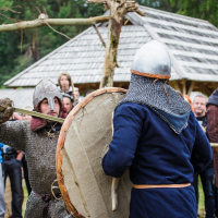 Two people in medieval combat attire engage in a mock sword fight during a reenactment event at Kilkim Žaibu 2015. They wear helmets and chainmail, and one holds a large round shield. Onlookers, some with cameras, observe the scene in front of rustic wooden structures set against a backdrop of trees.