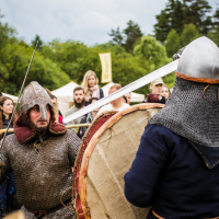 A medieval reenactment scene at the 2015 Kilkim Žaibu festival features two men in chainmail and helmets engaged in mock combat. One man holds a large round shield while the other thrusts a sword towards him. The background includes onlookers, tents, trees, and a cloudy sky, creating a lively historical atmosphere.