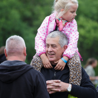 A gray-haired man smiles as he carries a young blonde girl on his shoulders. The girl is wearing a pink polka-dot jacket and leopard print pants. They are outdoors at the 2015 Kilkim Žaibu festival, with green trees visible in the background. Another man with short hair, facing away, is speaking to the man carrying the girl.
