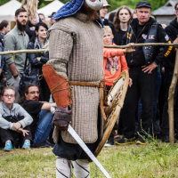A person dressed in medieval armor, including a helmet with a face guard, padded tunic, and leather gauntlets, stands with a sword and wooden shield. Spectators at the 2015 Kilkim Žaibu festival watch attentively in the background, suggesting this scene is part of a historic reenactment.