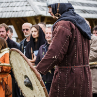A person dressed in medieval clothing and a metal helmet stands holding a large, round shield. The individual is garbed in a brown tunic with a cloth hood and belt. In the background, attendees of Kilkim Žaibu 2015, clad in both period attire and modern clothes, gather near a wooden structure with a shingled roof.