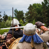Two people in medieval combat attire are engaged in a sword fight during the Kilkim Žaibu 2015 reenactment event. Both wear helmets and padded clothing. One carries a large round wooden shield, while the other wields a raised sword. A crowd of onlookers and trees are visible in the background.