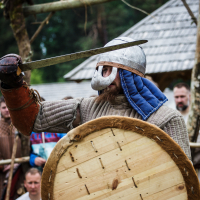 A person dressed in historical warrior attire participates in a reenactment at Kilkim Žaibu 2015. They wear a metal helmet with a blue fabric drape, chainmail, and a padded coat. Holding a round wooden shield in one hand and a drawn sword in the other, they perform as spectators watch the event unfold in the background.