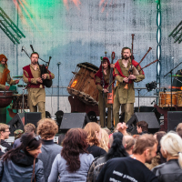 At Kilkim Žaibu 2015, a band performs on a stage at an outdoor event. The group features five musicians playing bagpipes alongside a drummer. Clad in medieval-inspired clothing in brown and red tones, they captivate the crowd gathered before the stage, illuminated by vibrant lighting and speakers.