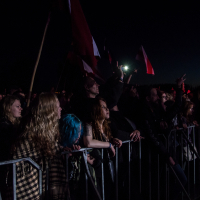 A crowd of people at an outdoor festival stands behind a metal barricade, looking towards the stage with intensity. Some hold flags, while others lean forward, engrossed in the Kilkim Žaibu performance. The image is dimly lit, reflecting a nighttime setting with a spotlight casting a red hue on some faces.
