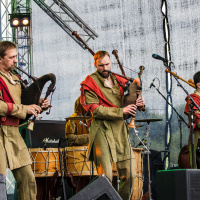 Three musicians on a stage during Kilkim Žaibu 2015 play bagpipes. They are dressed in traditional, earthy-toned outfits accented with red fabric draped around their shoulders. The background features stage equipment and a drum set, with trees faintly visible beyond the stage. Mic stands are scattered around.