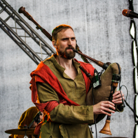 At the 2015 Kilkim Žaibu festival, a man in a green tunic and red cloak plays the bagpipes on stage with a serious expression and a trimmed beard. The background reveals stage lighting and equipment, while another person in similar attire is partially visible, playing a drum set.