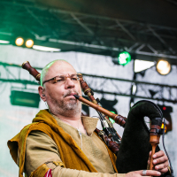 A man with glasses and a bald head is playing a bagpipe on stage during the 2015 Kilkim Žaibu festival. He is wearing a medieval-style outfit consisting of a brown tunic with green accents. The stage behind him is lit with green lights, and a truss with additional lights is visible in the background.