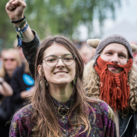 A young woman with long brown hair, glasses, and a patterned shirt stands smiling outdoors. Behind her, a person with a knitted hat resembling a Viking helmet and red beard raises a clenched fist. Blurry greenery and more people are in the background, indicating the lively atmosphere of Kilkim Žaibu 2015.