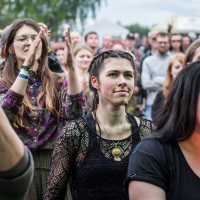 A crowd of young adults at an outdoor event, capturing the lively and engaged mood of Kilkim Žaibu 2015. One woman in the foreground has braids and wears a black netted top with a circular pendant. Another person is clapping. Trees and a white tent are visible in the background.