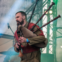 A bearded man plays the bagpipes on a stage at the 2015 Kilkim Žaibu festival, wearing a red and brown outfit. Behind him are stage lights and structural rigging. A blue wristband is visible on his right wrist. The stage is filled with smoke, and the drummer in the background is partially visible. Image credit "xltphoto.net" is in the bottom