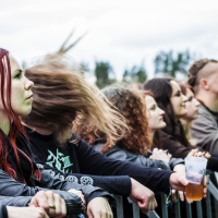 People stand behind a metal barricade at Kilkim Žaibu 2015, an outdoor concert. In the foreground, a woman with long red hair faces the stage, and a person next to her has their hair flying wildly. Many attendees focus on the performance, with one individual holding a beer cup. Trees blur in the background.
