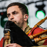 A man with short hair and a trimmed beard plays a bagpipe on stage at Kilkim Žaibu 2015. He wears in-ear monitors and is dressed in a green outfit with an orange-red shawl over his shoulder, performing amidst blurred lights and equipment in the background.