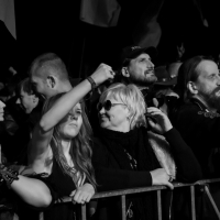 A black and white image captures the energy of the Kilkim Žaibu festival in 2015. The outdoor concert crowd is tightly packed at night, with some hands raised. The central figure, wearing sunglasses, smiles and looks right. Another person with a beard and long hair stands to the right, as flags wave in the background.